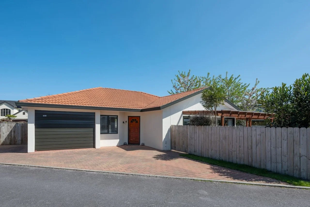 A single-story house with a red tiled roof, white exterior walls, a black garage door, a wooden front door, a driveway, a wooden fence, and some trees and shrubs in the yard under a clear blue sky.