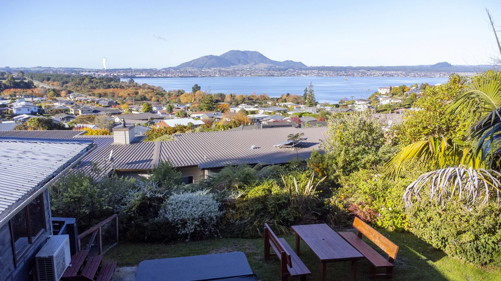 View of a residential neighborhood near a large body of water with mountains in the background, seen from a backyard with outdoor furniture and lush greenery.
