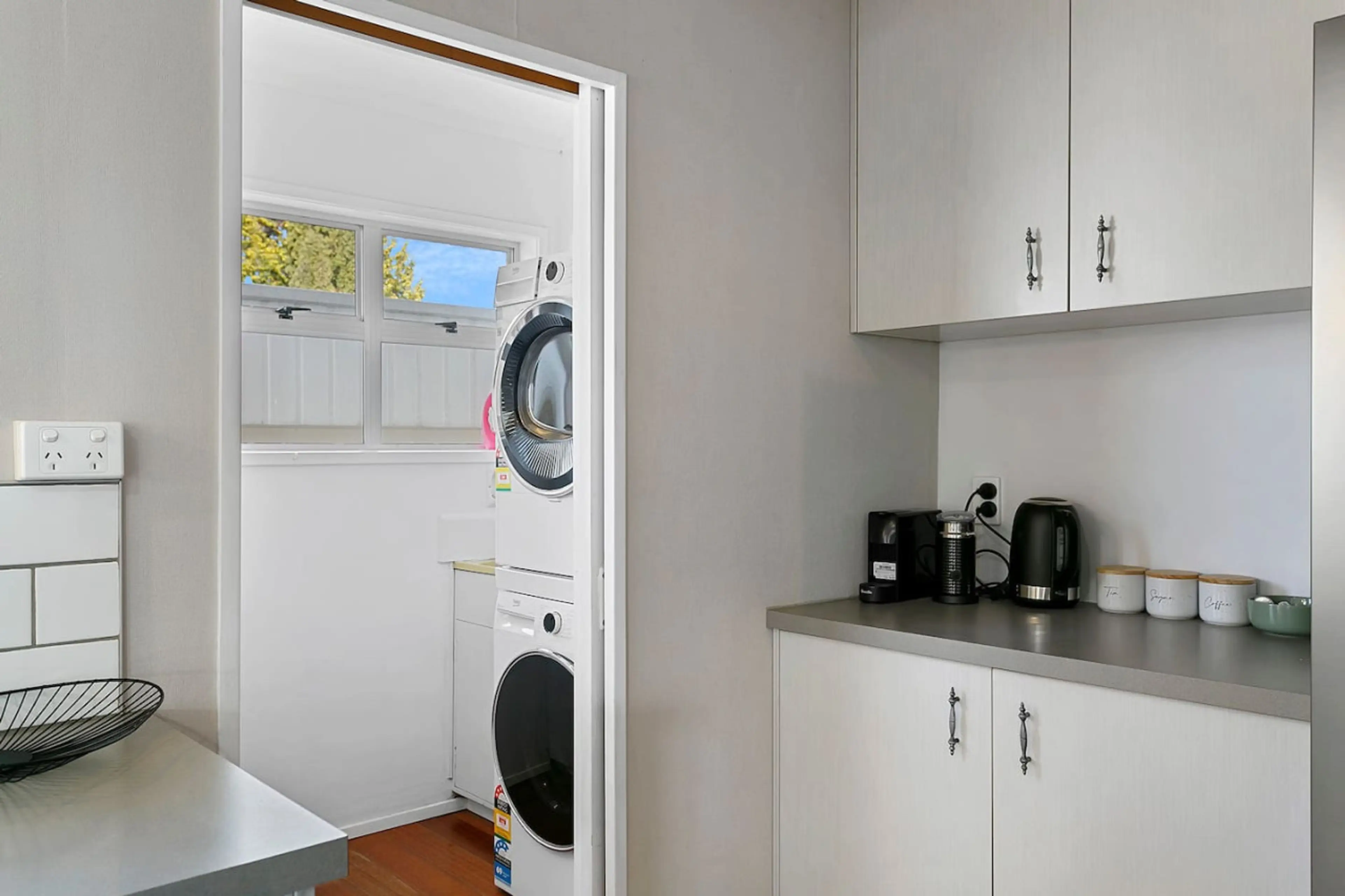 Laundry room with stacked washer and dryer, window with trees outside, countertop with coffee machine, kettle, and canisters labeled Tea, Sugar, Coffee.