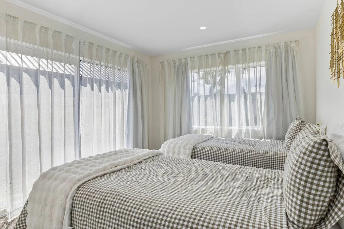 Bedroom with two single beds, checkered bedding, and sheer curtains covering large windows, allowing natural light to fill the room.