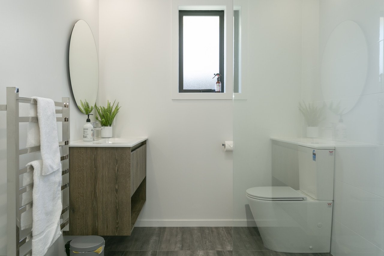 Modern bathroom with white walls, a small window, a wooden vanity with a white countertop, a potted green plant, an oval mirror, a towel heater, and a white toilet.