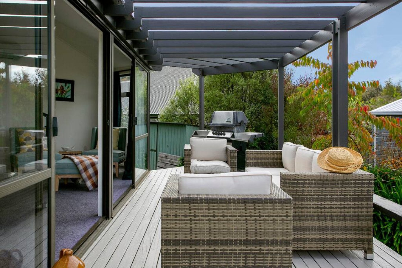 Outdoor patio with wicker furniture, a gas grill, and surrounding greenery, attached to a house with sliding glass doors leading to an indoor room.