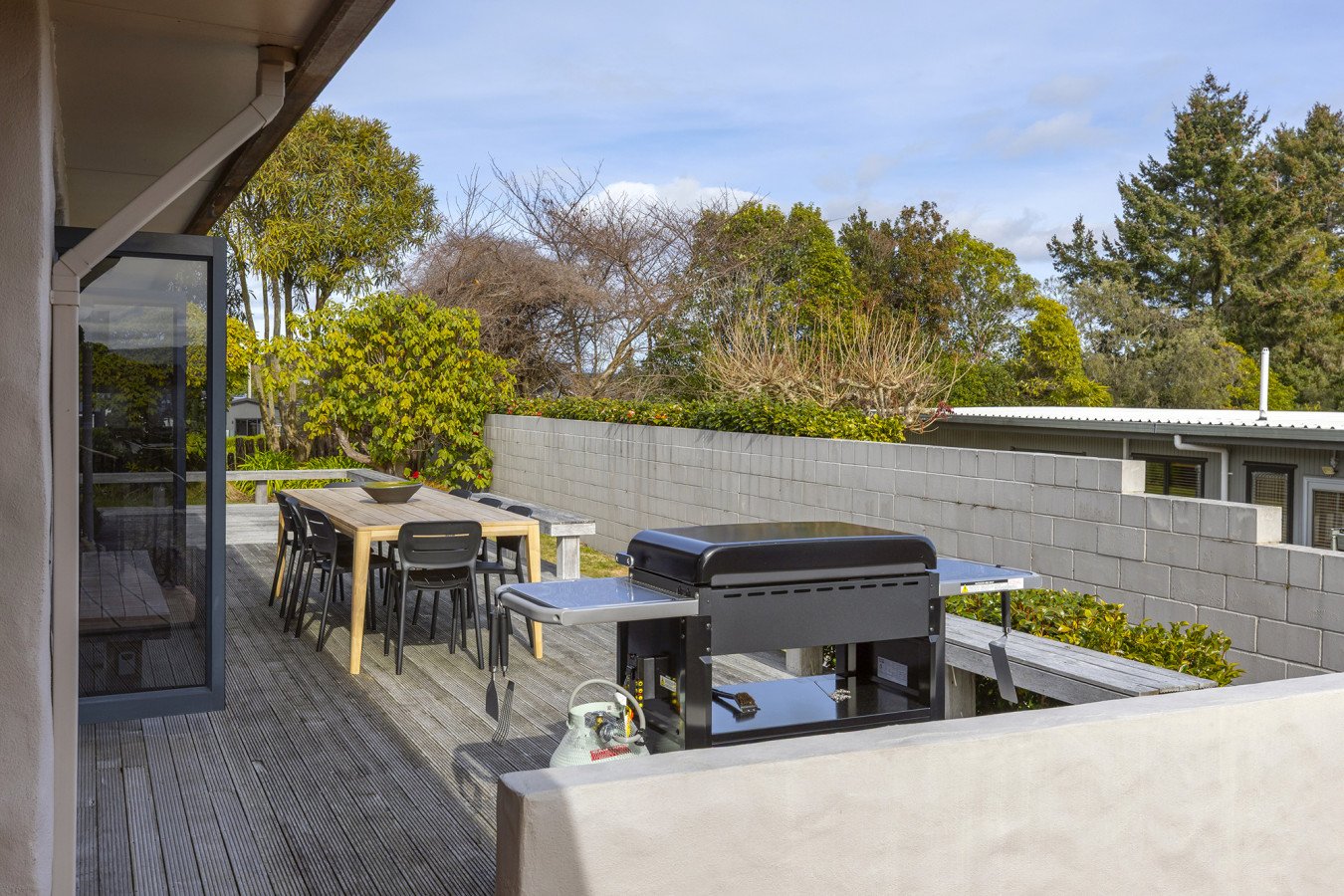 Outdoor patio with wooden dining table, black chairs, and a grill, surrounded by trees and a concrete block wall.