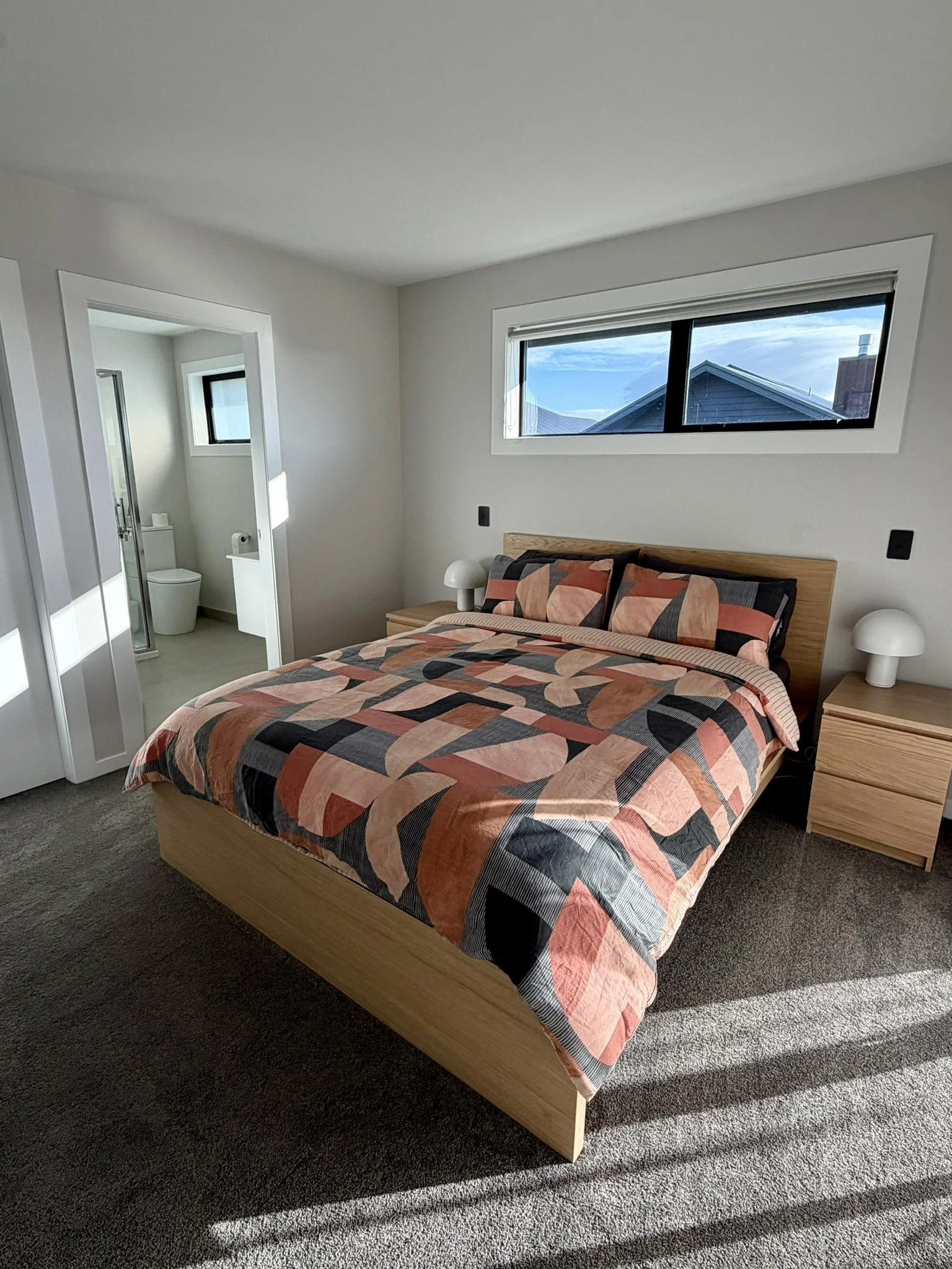 Modern bedroom with wooden bed frame, patterned bedding, two nightstands with lamps, and a window showing rooftops outside. Bathroom with toilet and small window visible through open door.