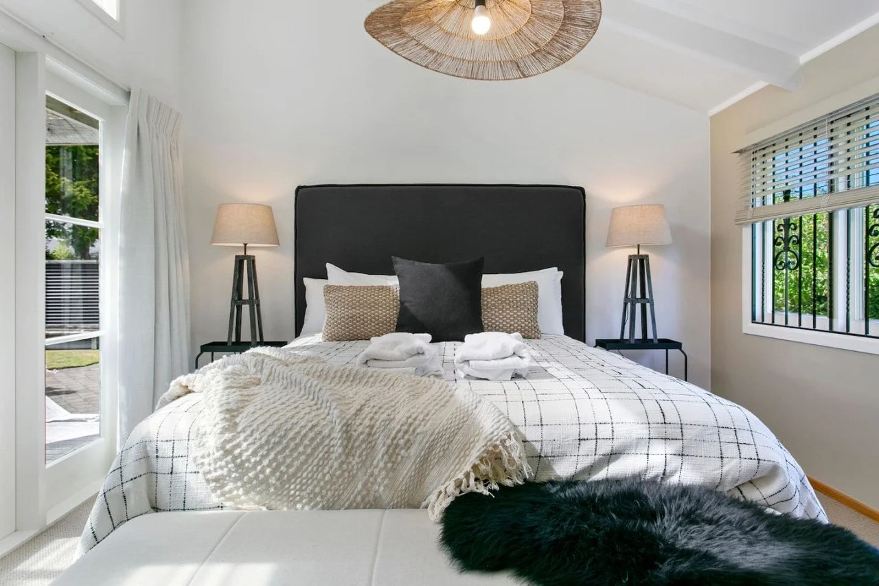 A neatly made bed with a black headboard, two beige and white pillows, a black decorative pillow, and a white plaid bedspread. Two bedside lamps on black nightstands, and a window with blinds and greenery outside. A cream throw blanket is draped over