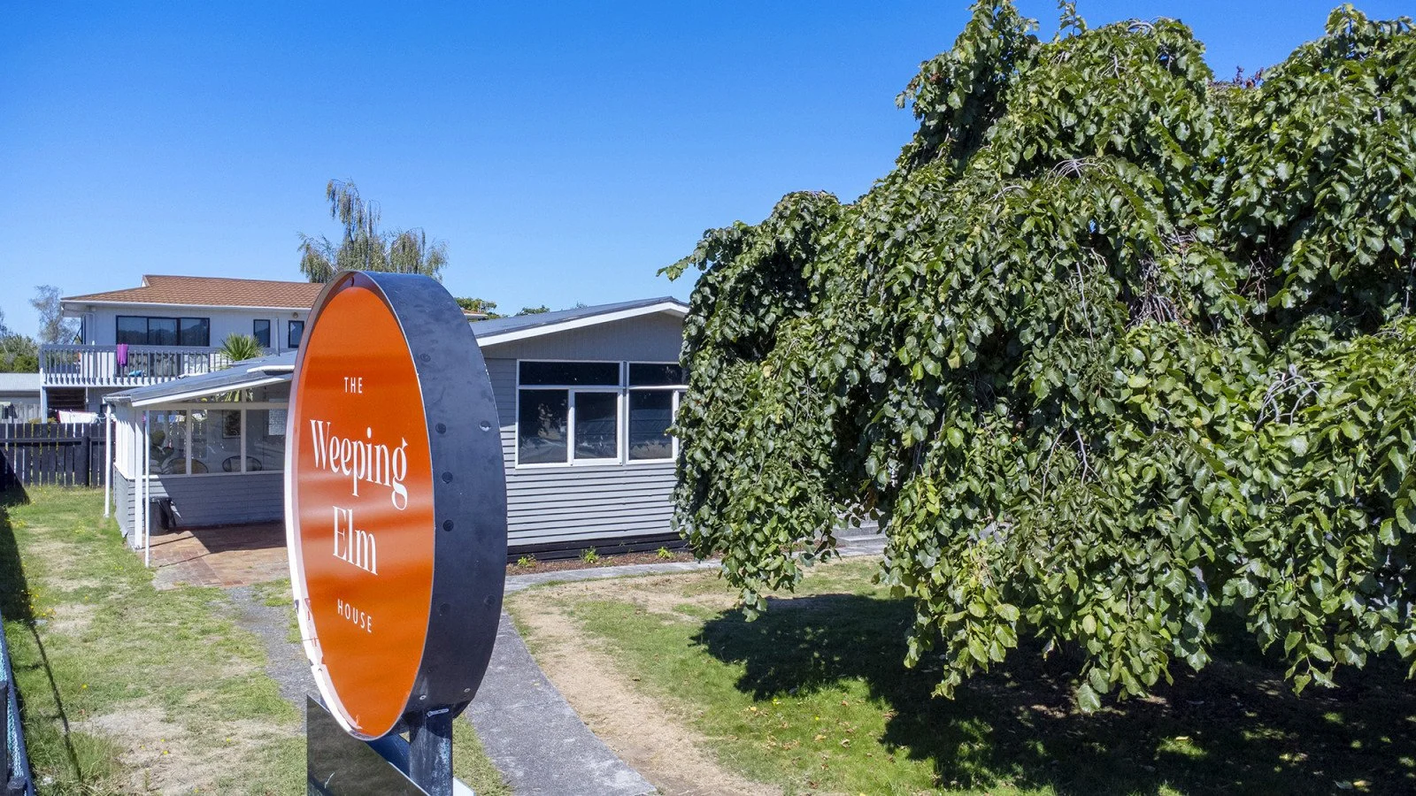 Sign for the Weeping Elm House near a house and large green tree in a sunny yard.