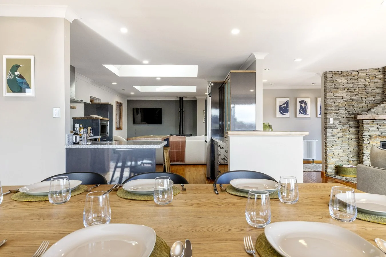 Dining area with a wooden table set with white plates, glasses, and cutlery, overlooking a modern open-concept living space with a stone fireplace, artwork, and a kitchen.