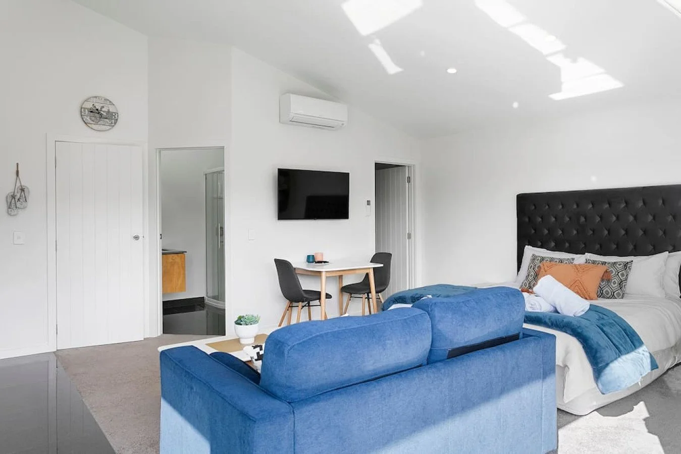 Modern bedroom with a bed, blue sofa, wall-mounted TV, small eating table with two chairs, and a flat white wall with minimal decorations.