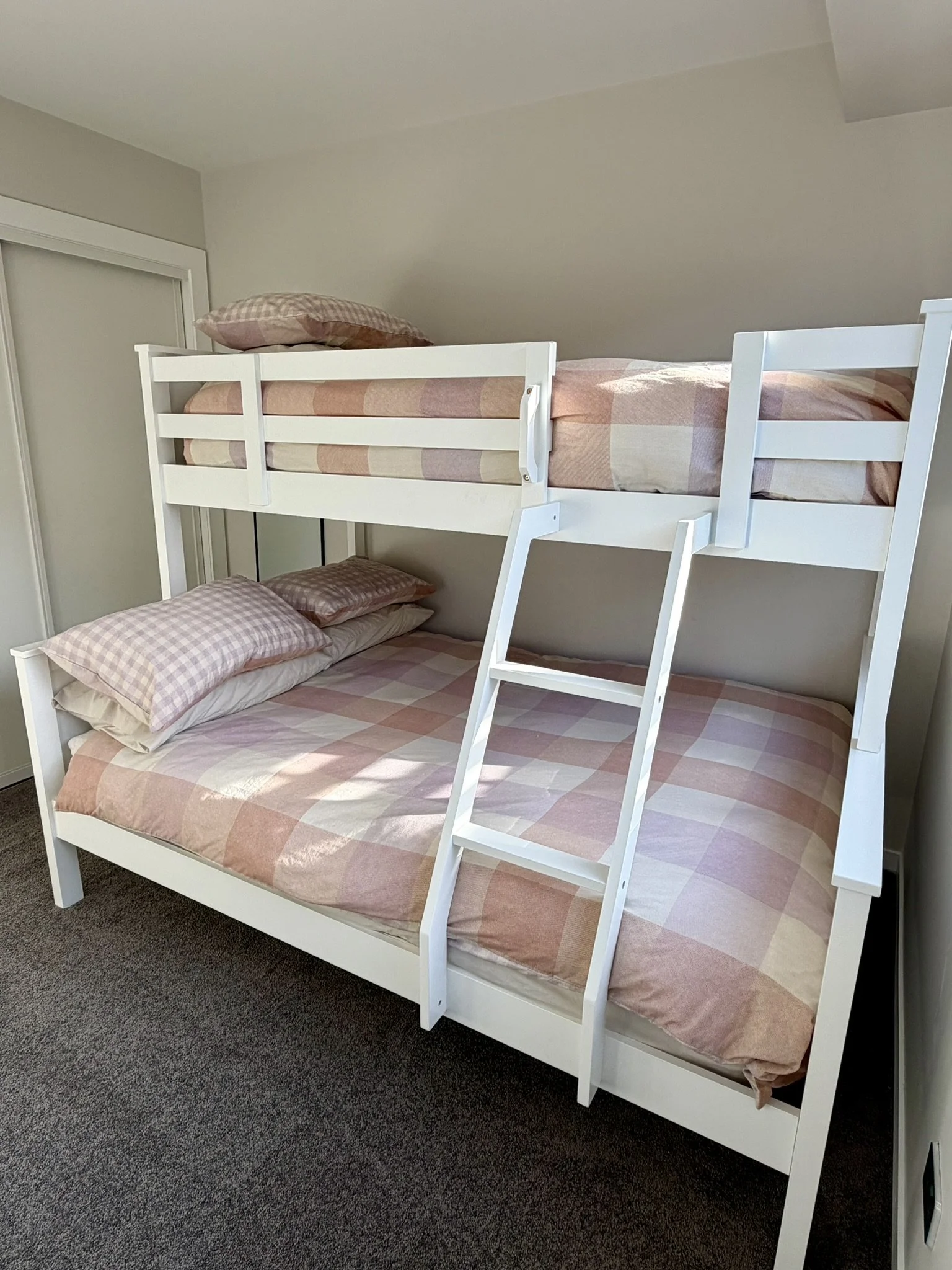 White bunk bed with pink gingham and plaid bedding, pillows, and a ladder, positioned against a light beige wall on a dark carpeted floor.