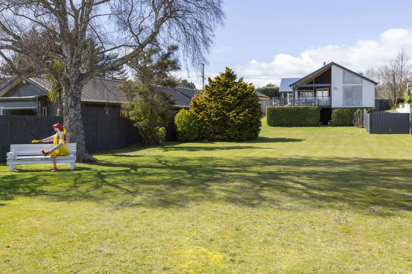 A backyard scene with a large tree, green grass, and houses in the background. A colorful clown statue with red hair, striped sleeves, and yellow dress is sitting on a white bench under the tree.