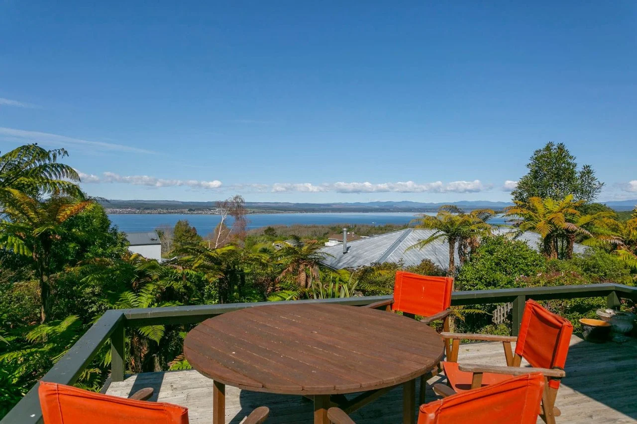 A wooden deck with four orange chairs around a round table. The deck overlooks lush greenery, a lake, and distant hills under a clear blue sky.