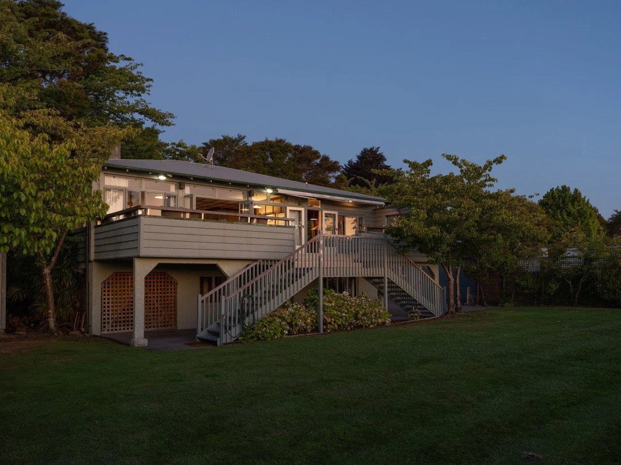 A two-story house with a large deck, stairs leading up to the deck, surrounded by trees and a lawn, with lights on and the sky in dusk.