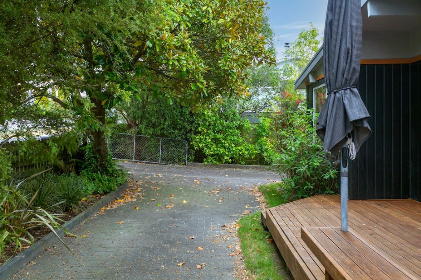 A backyard with a paved pathway, green trees, and a small deck with a black umbrella.