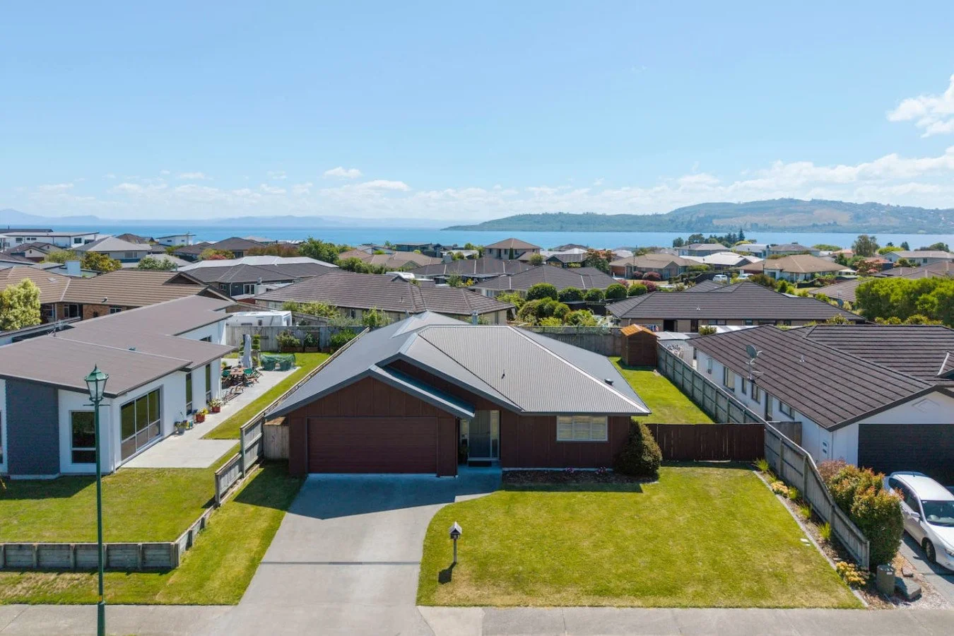 Aerial view of a suburban neighborhood with single-story houses, green lawns, and a lake in the background under a blue sky.