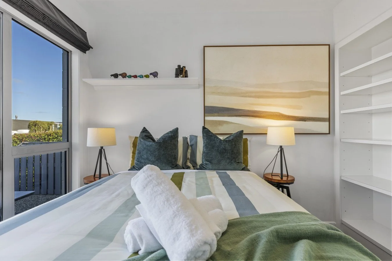 Bedroom with a bed, dark green pillows, bedside tables with lamps, window showing outdoor view, and a large abstract landscape painting above the bed.