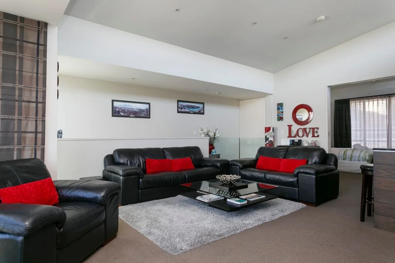 Living room with black leather sofas, red pillows, grey rug, glass coffee table, wall decor including a red 'LOVE' sign, framed pictures, a small side table with flowers, and a window with curtains revealing a seating area with cushions.