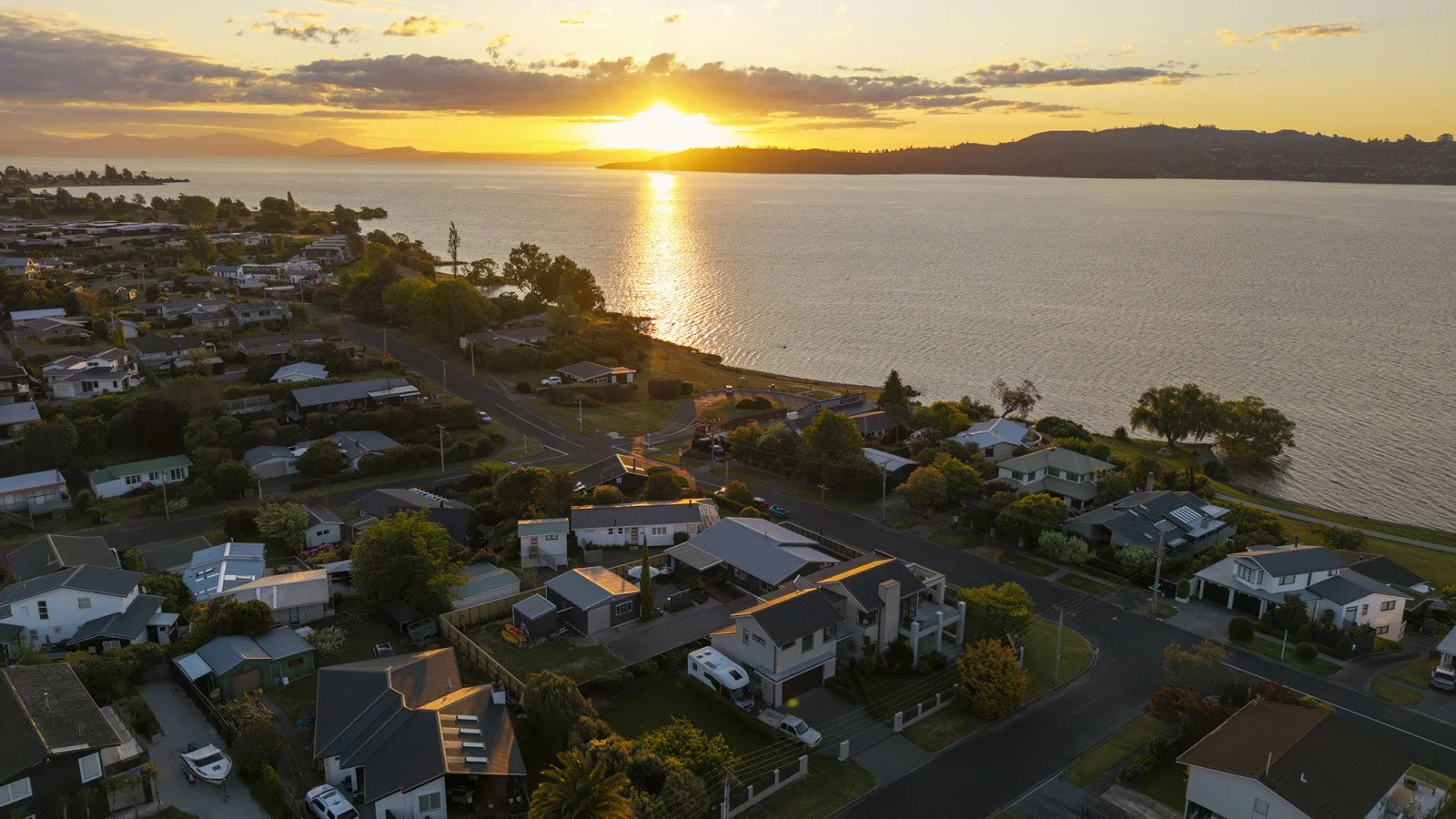 An aerial view of a coastal residential neighborhood during sunset, featuring houses, trees, and roads along a shoreline with a large body of water and hills in the distance.