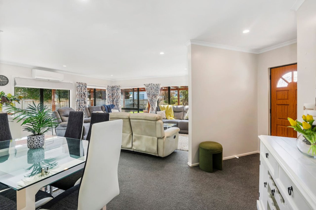Open living and dining room area with beige and gray sofas, dining table with chairs, large windows with floral curtains, and a white sideboard with yellow flowers.