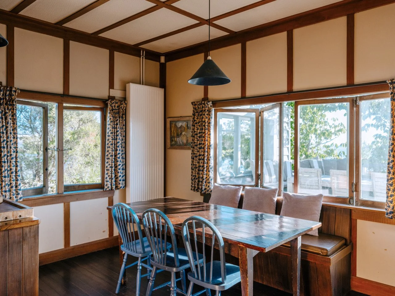 Dining room with a rustic wooden table surrounded by six chairs, three cushioned bench seats, large windows, floral curtains, and a pendant light fixture hanging from the ceiling.