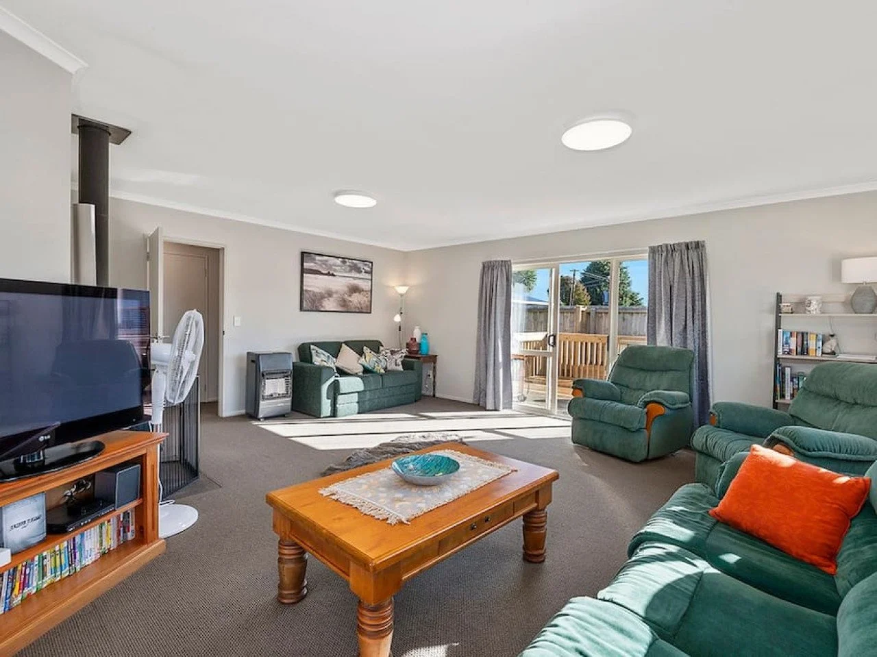 Living room with green sofas, a wooden coffee table, a TV on a wooden stand, sliding glass door leading to a deck, and a bookshelf.