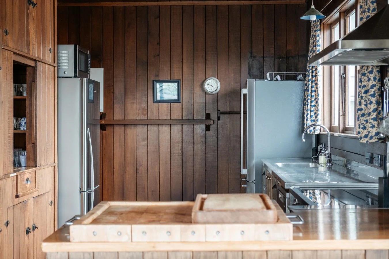 Kitchen with wooden wall panels, a refrigerator, a microwave on top, a small picture frame, a round clock, a stainless steel sink under a window with floral curtains, and a wooden countertop with a cutting board.