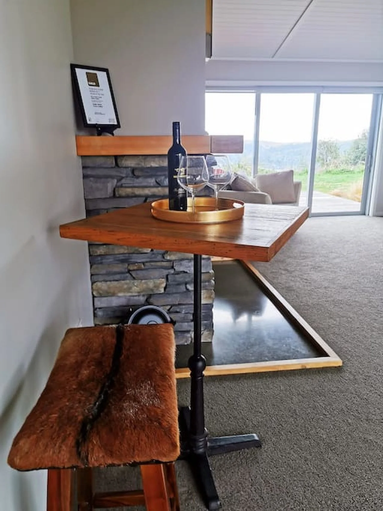 A cozy living space with a small wooden table featuring a bottle of wine, two glasses, and a round tray. There is a furry patchwork stool in the foreground and a stone fireplace behind the table. Large windows in the background reveal a scenic outdoo