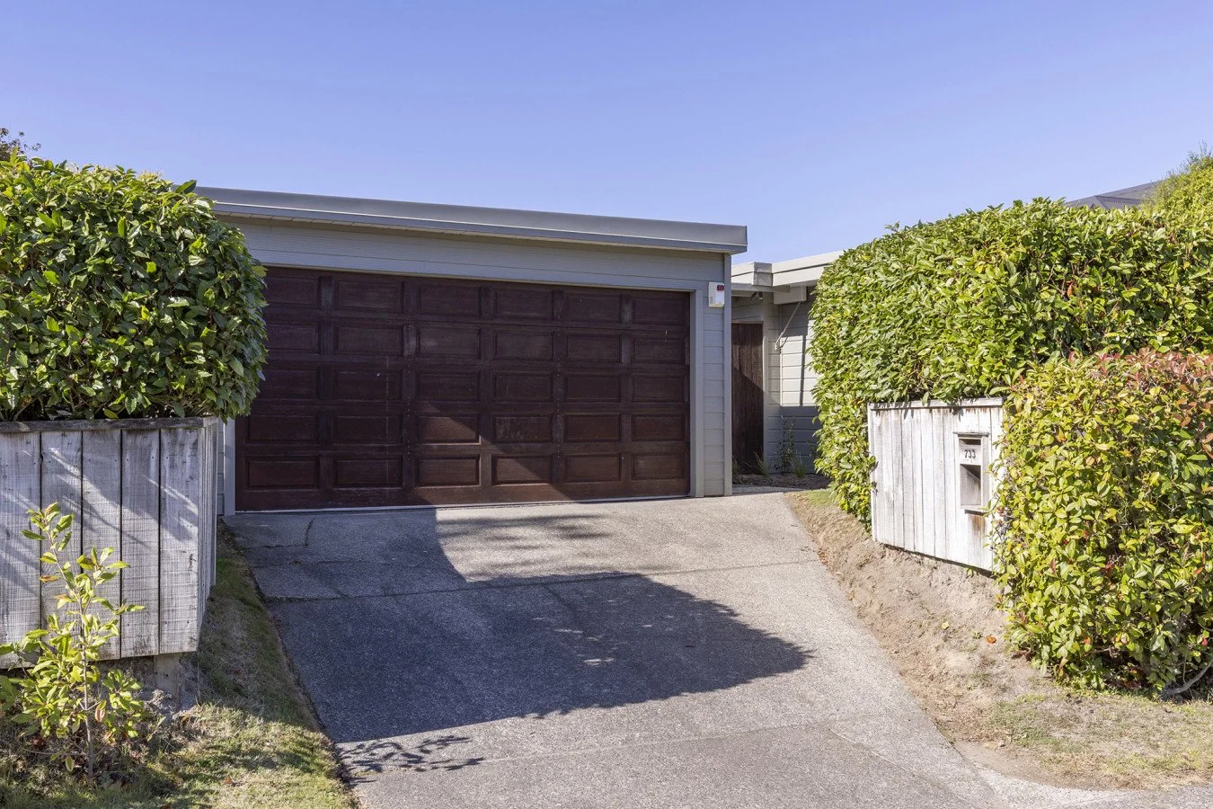 Residential house with a brown garage door, driveway, and green bushes on both sides, under a clear blue sky.