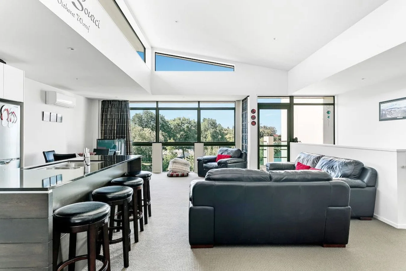 Modern living room with large windows, black leather sofas, a dining table with chairs, and a view of trees outside.