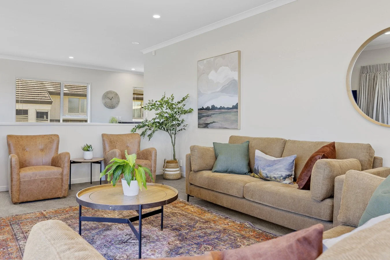 Living room with beige sofa, armchairs, potted plants, framed artwork, round mirror, and window blinds.