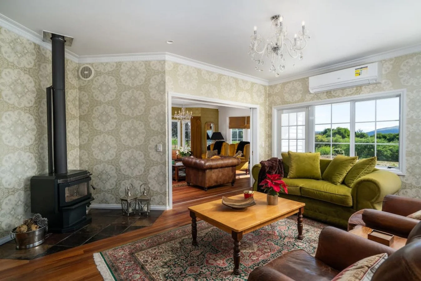 Living room with green sofa, brown armchairs, wooden coffee table with a poinsettia plant, patterned area rug, large window showing a green landscape, wood stove, brick floor section, wallpaper with a white and beige pattern, chandelier, air conditio