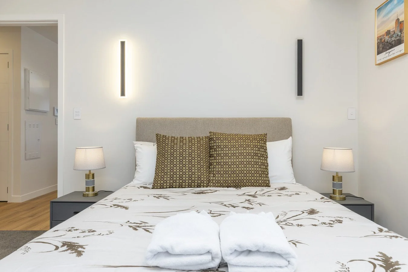 Modern bedroom with beige upholstered headboard, two decorative pillows, white bedding with brown floral pattern, two bedside lamps, abstract wall art, and clean, minimalist decor.