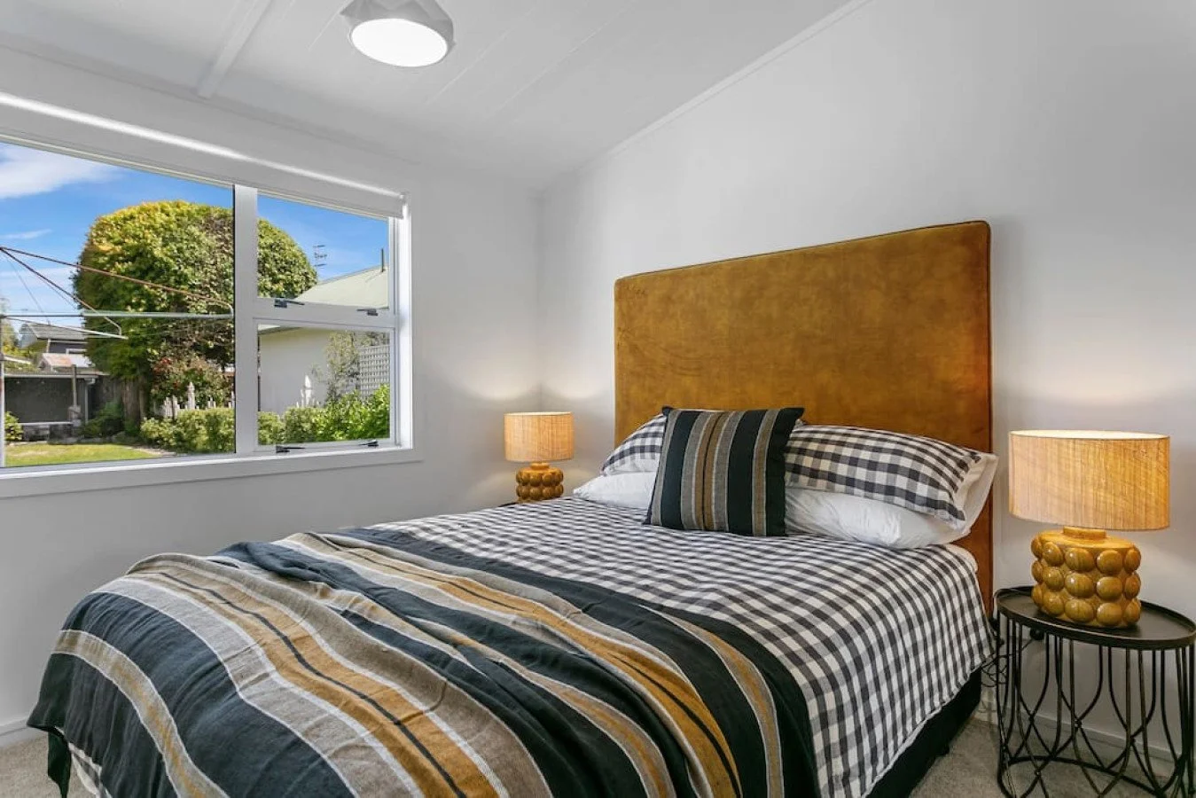 Bedroom with a bed featuring a checkered black, white, and yellow bedspread, striped pillow, and wooden headboard. There are two lamps on side tables and a window showing a garden outside.