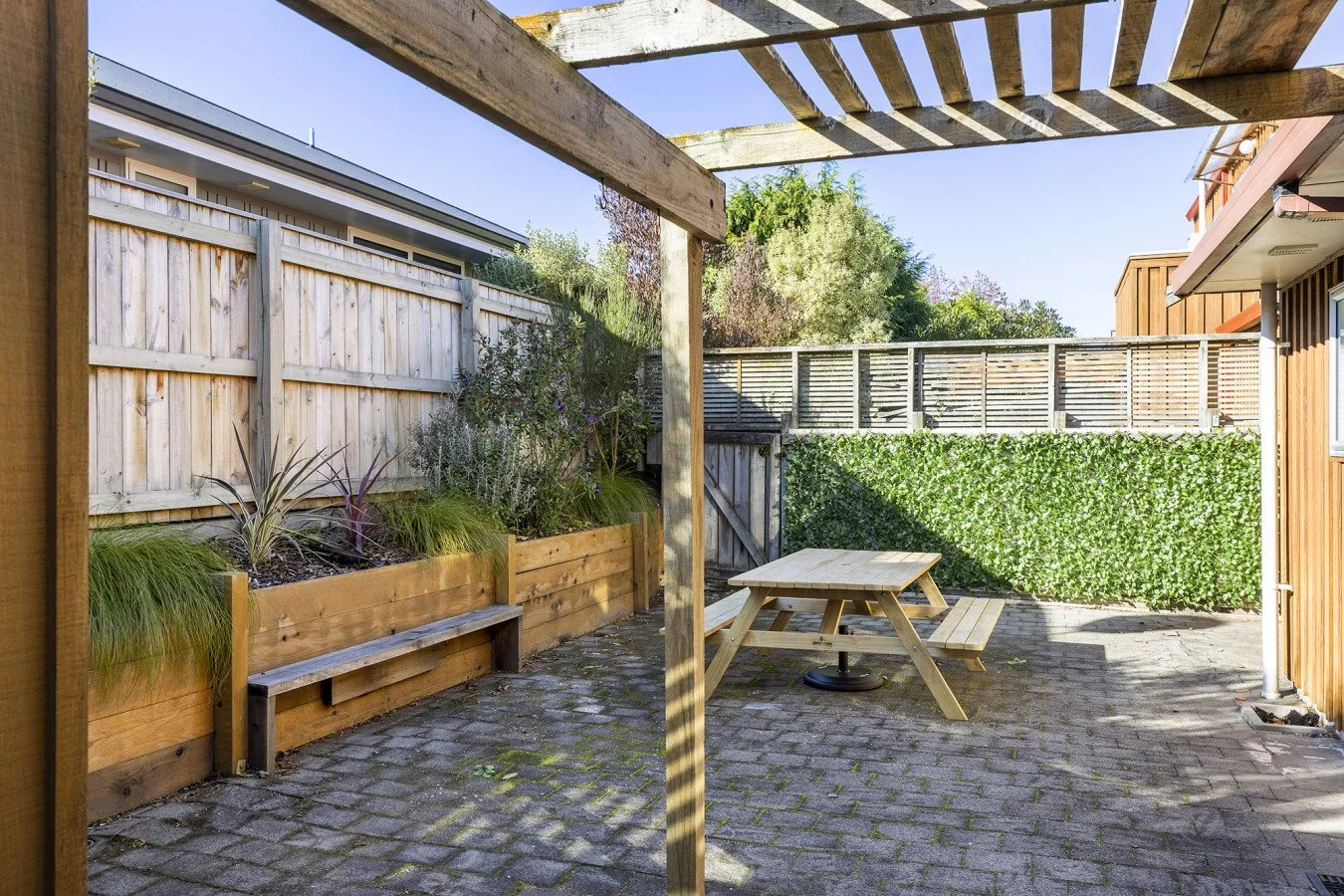 View of a backyard patio with a wooden table and bench, potted plants on the left, a wooden fence, and a hedge, under a pergola with shadows cast on the cobblestone ground.