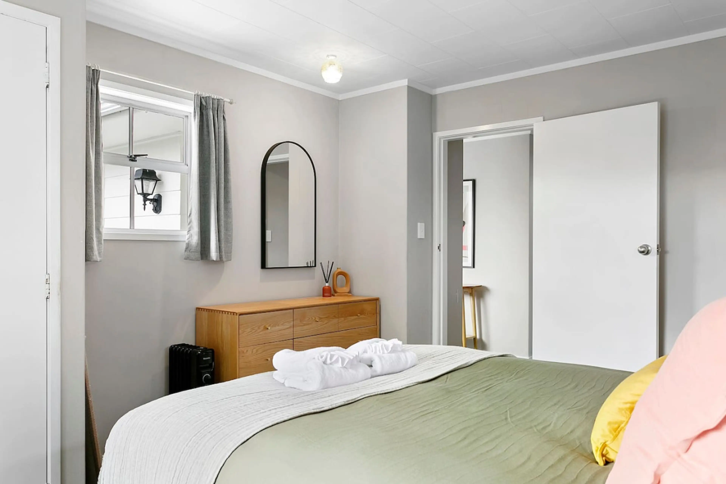 A neatly made bed with white, green, and pink bedding, two folded towels on top, a wooden dresser with a mirror, and a window with grey curtains in a minimalist bedroom with light grey walls.