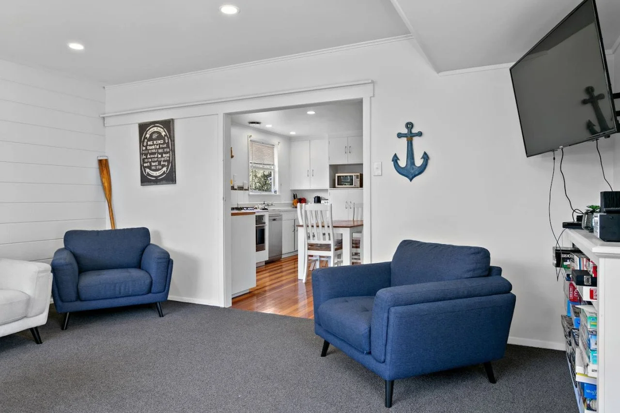 Living room with two blue armchairs, a wall-mounted television, and a white bookshelf with various items. The room opens to a kitchen with white cabinets and a wooden dining table. Nautical-themed decor includes a blue anchor and a black sign on the 