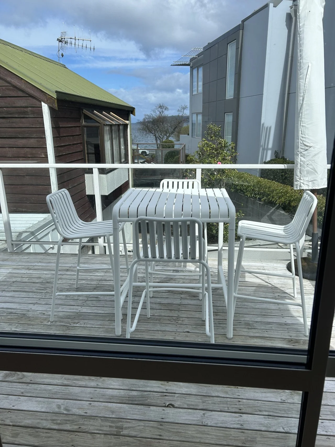 View of an outdoor balcony with white patio furniture and a glass railing, overlooking neighboring buildings and a cloudy sky.
