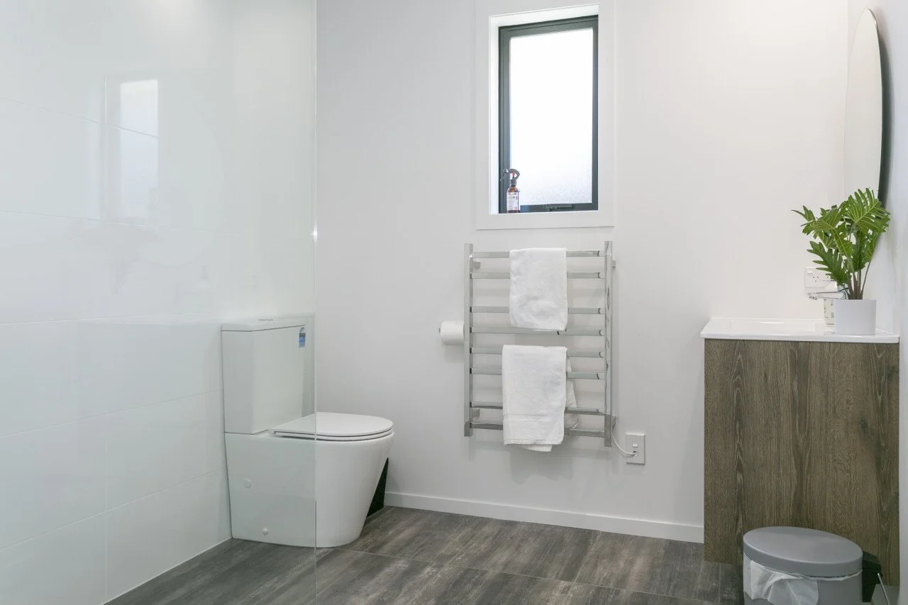Modern bathroom with white walls, a gray window, a toilet, a towel rack with white towels, a small cabinet with a plant, a mirror, and a trash can.