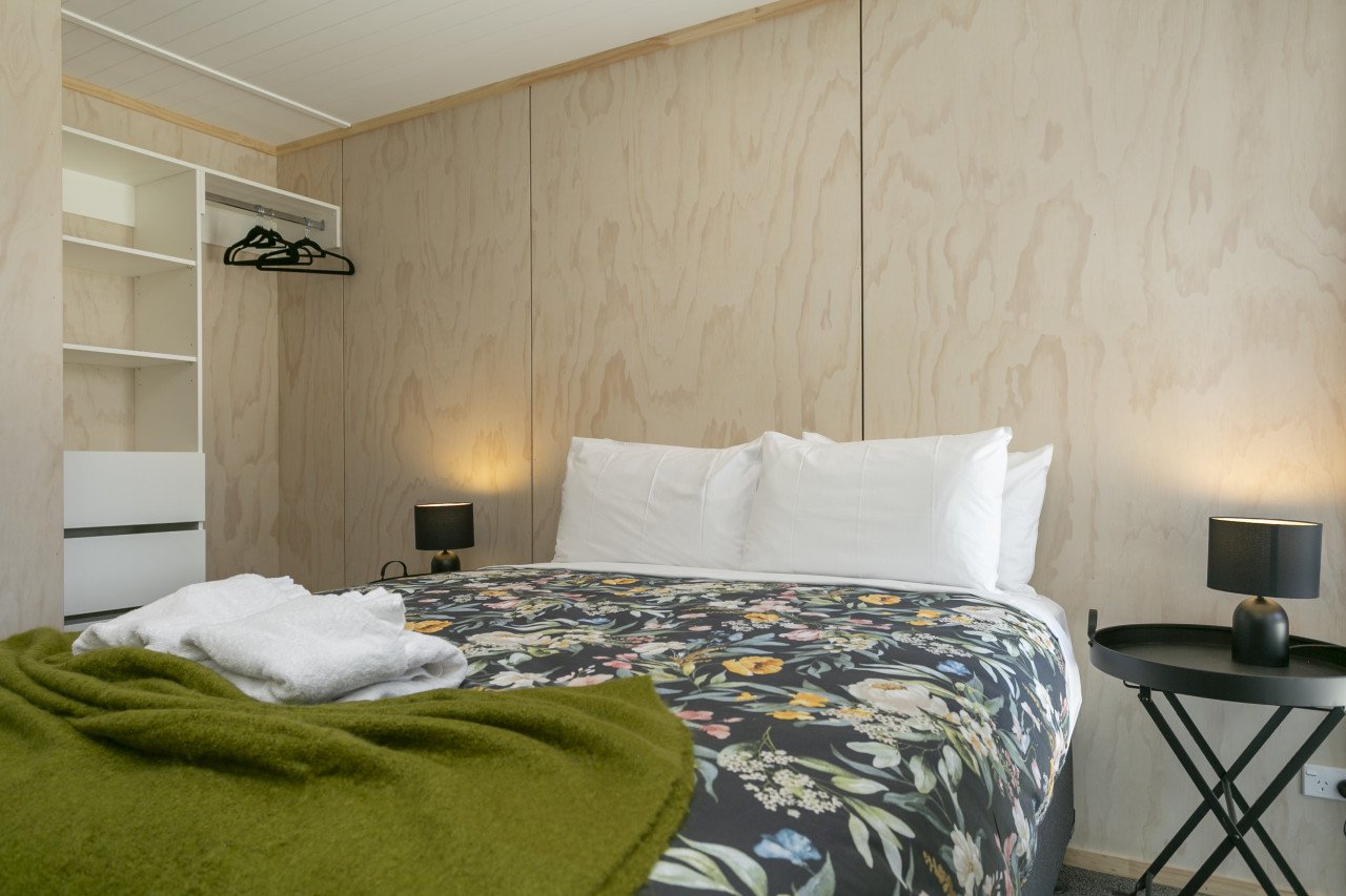 A neatly made bed with white pillows and a floral bedspread in a modern bedroom. There are two black bedside lamps, a small black tray table, and an open closet with hanging space and shelves in the background.