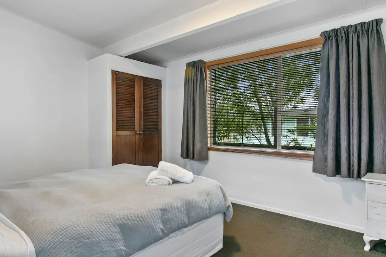 A bedroom with a bed, white sheets, a rolled towel on the bed, dark wood closet doors, a window with blinds, and curtains showing a view of trees and neighboring houses outside.