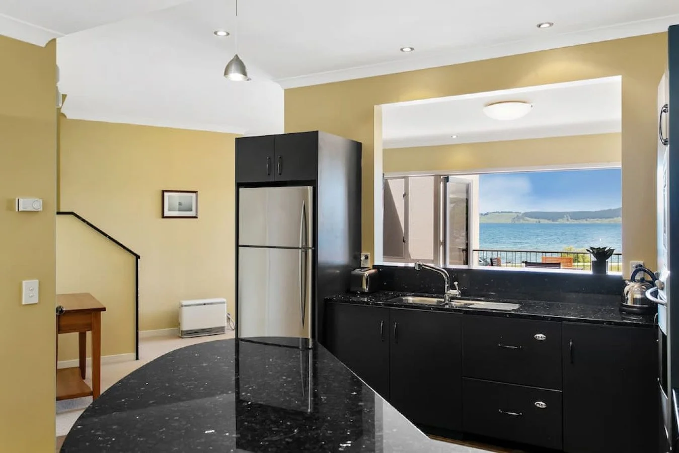 Modern kitchen with black cabinets, a stainless steel refrigerator, black granite countertops, and large windows showing a view of the ocean.