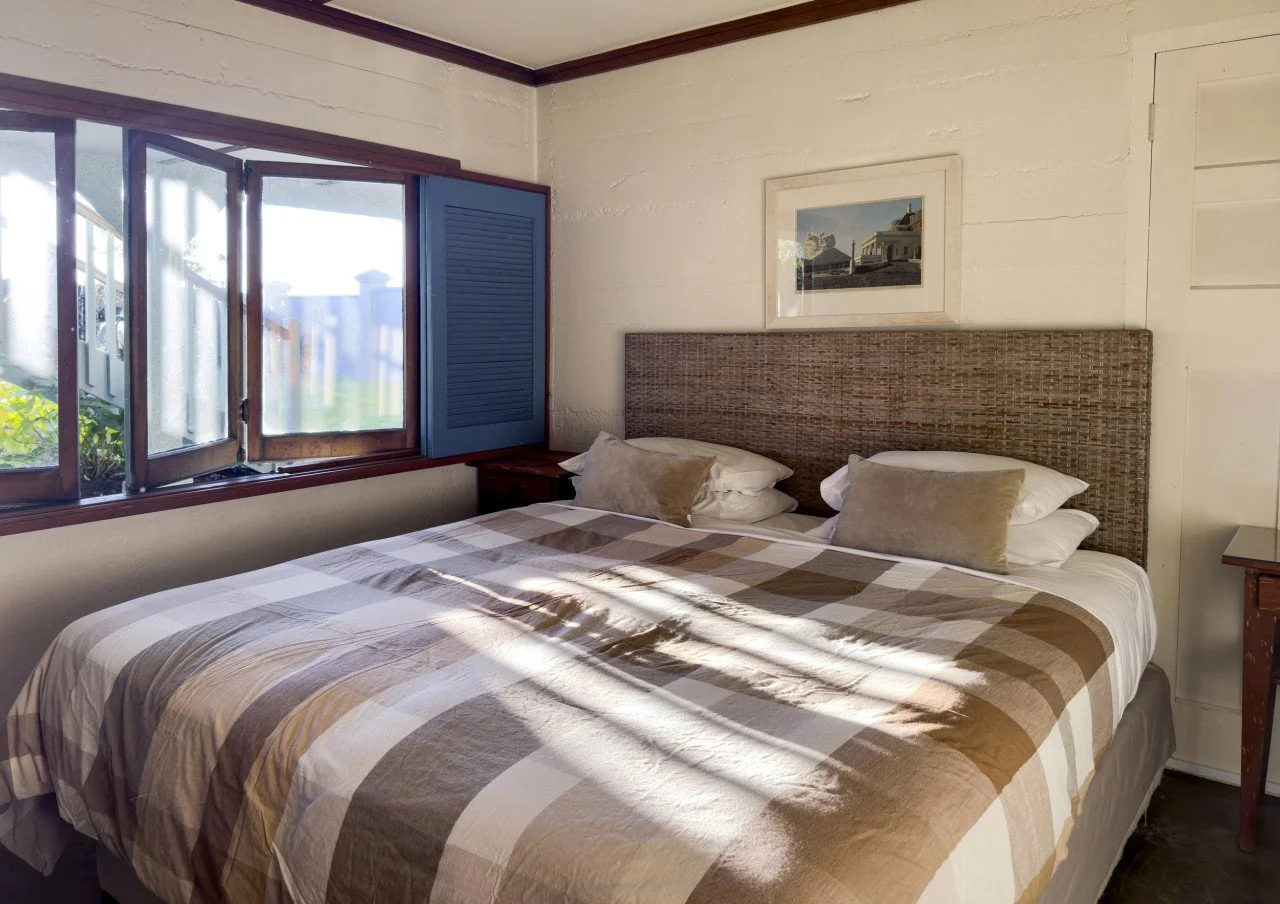 A cozy bedroom with a large bed featuring a plaid beige and brown bedspread and four pillows. There is a window with open wooden shutters letting in sunlight, and a framed picture hanging above the bed's headboard. The room has white painted wooden w