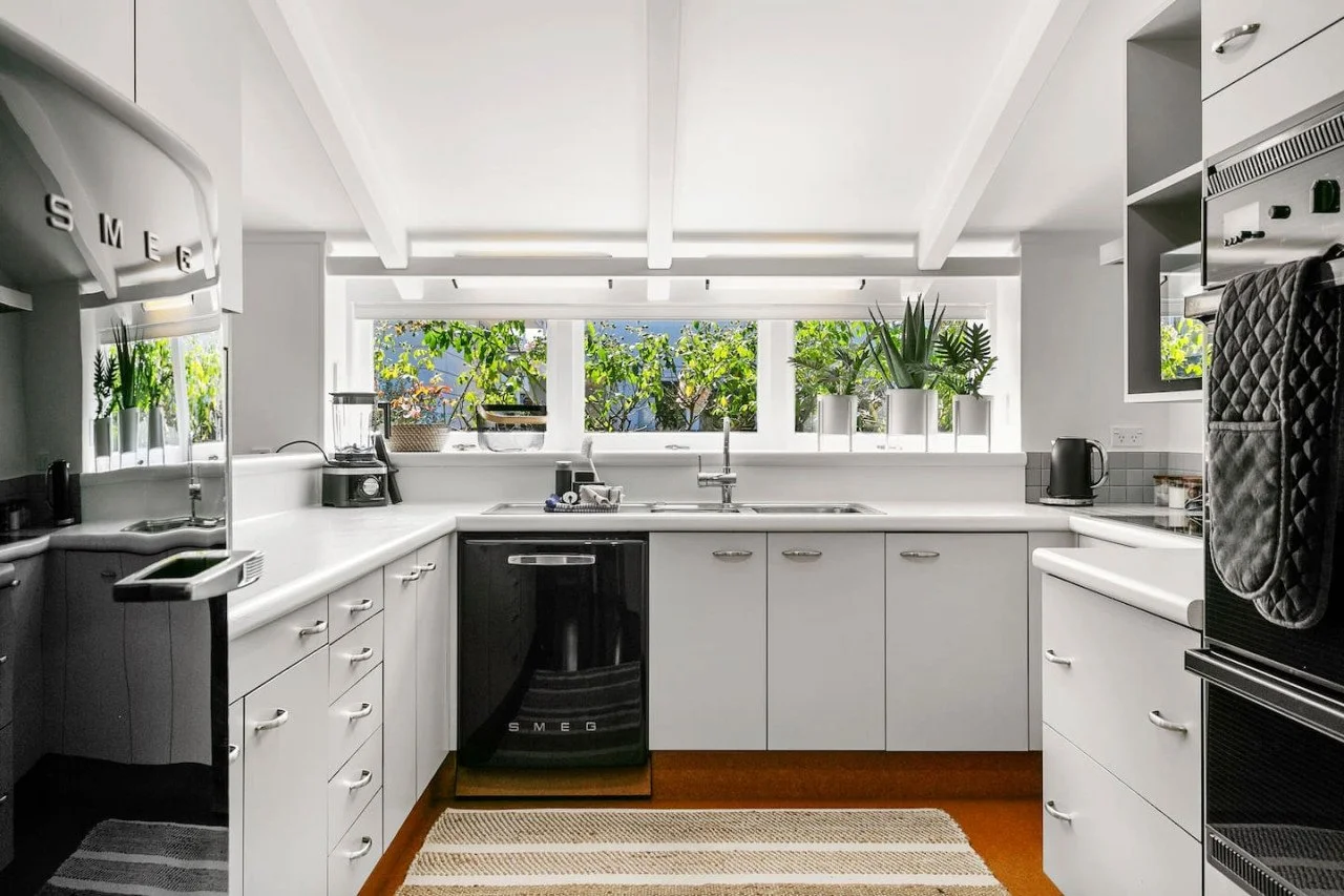 Bright kitchen with white cabinets, black SMEG dishwasher, and large window with plants outside.