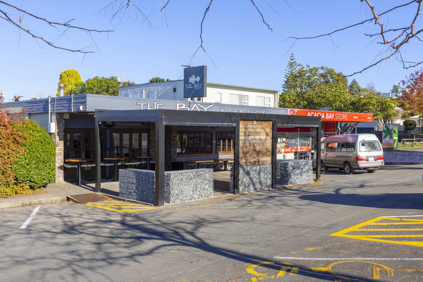 Exterior of a restaurant called 'The Bay Bar & Grille' with outdoor seating, a parked minivan, and trees in the background.