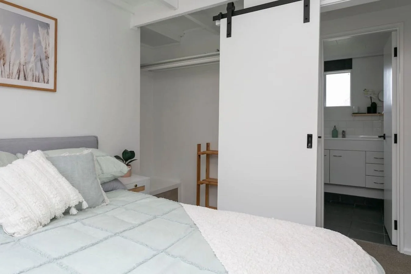 A bedroom with a bed, pillows, a nightstand with a plant, and a sliding closet door. An open door reveals a bathroom with a vanity and a window.