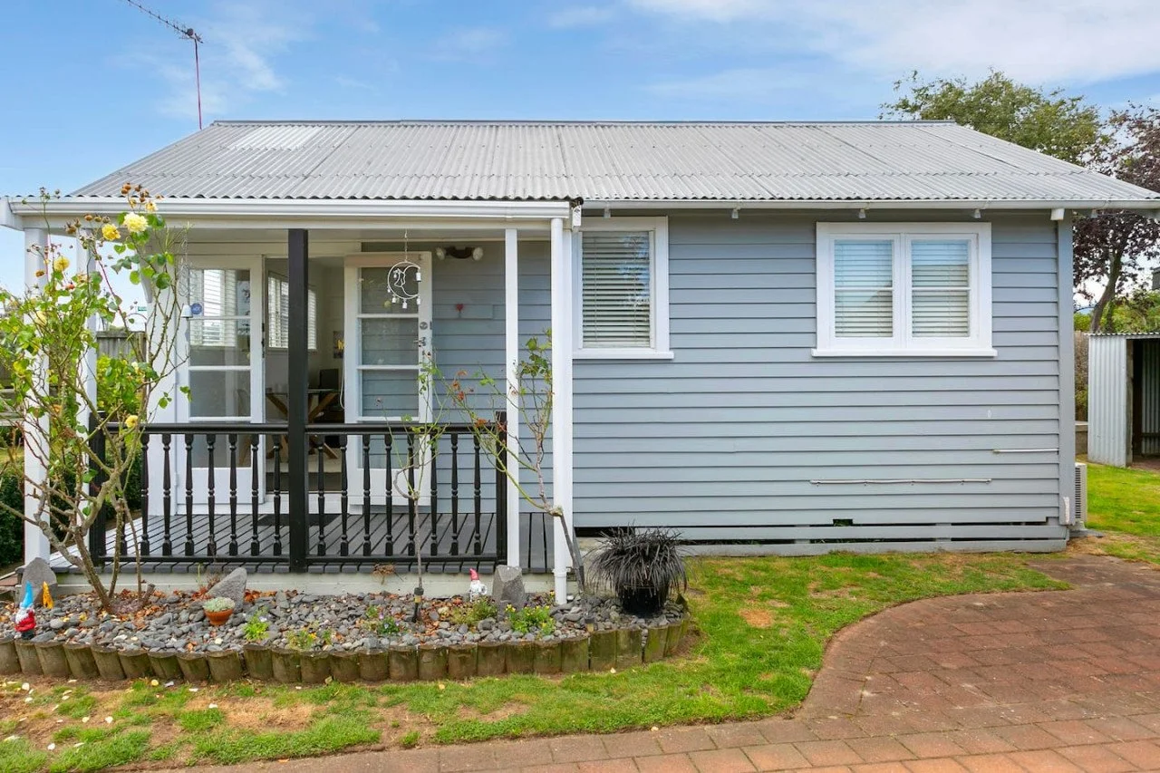 Front view of a light blue house with white window frames and a gray corrugated metal roof. There is a small front porch with black railing and a garden bed with rocks and plants in front. A curved brick pathway is visible on the right side.
