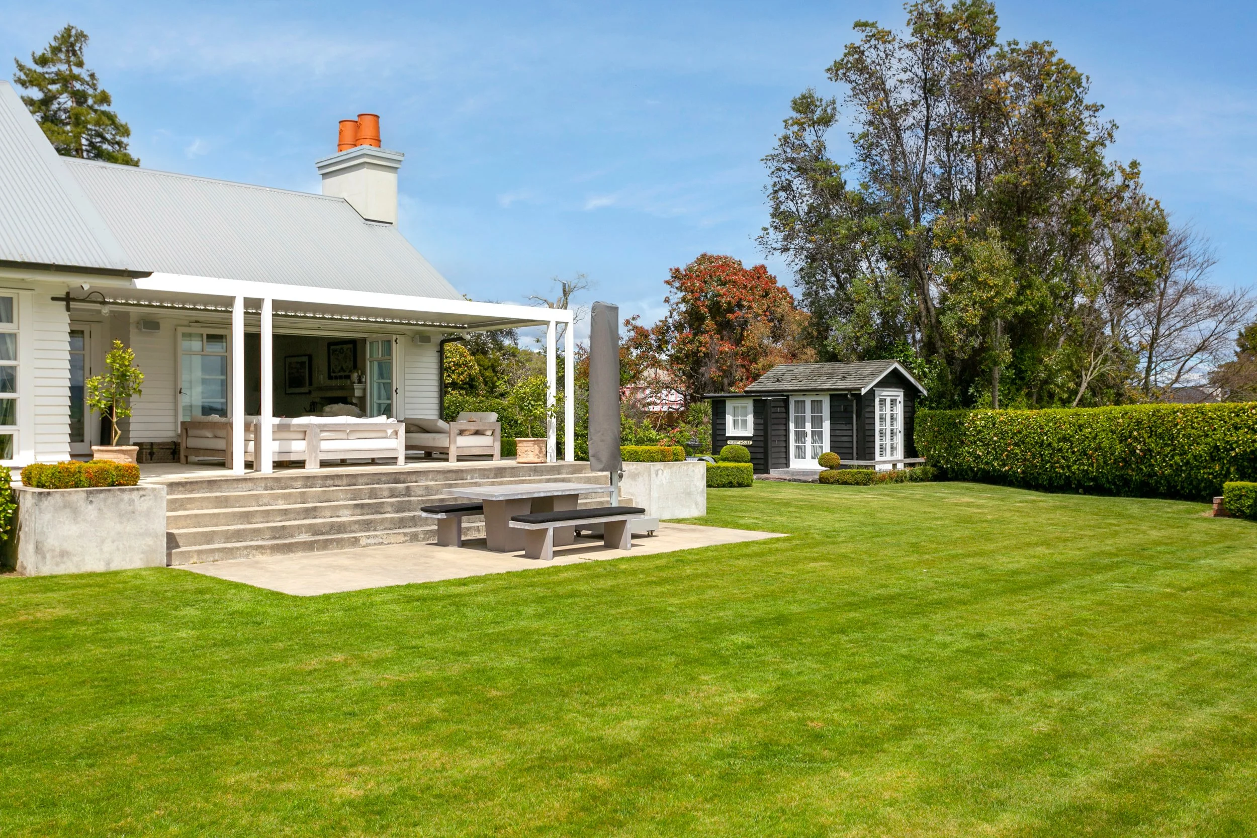 A backyard with a white house, a concrete patio with a picnic table, a small black shed, well-maintained grass, bushes, trees, and a partly cloudy sky.