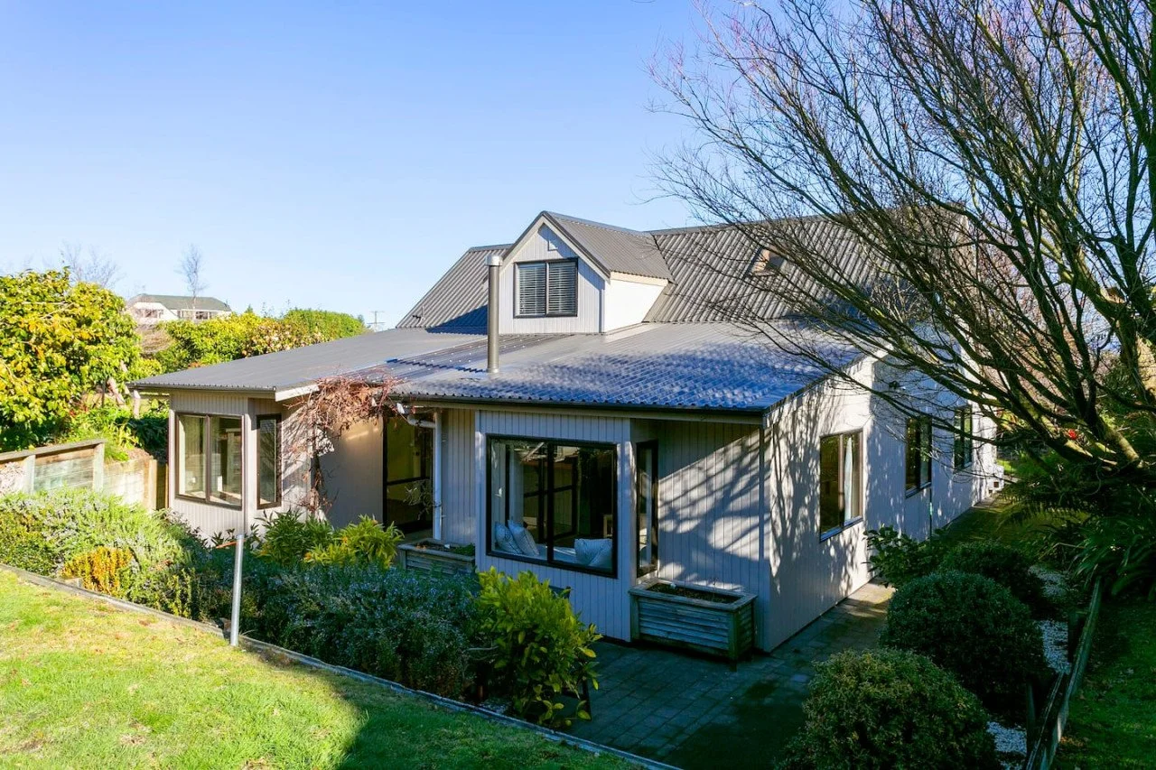 A two-story house with a metal roof, surrounded by trees and bushes, with clear blue sky in the background.
