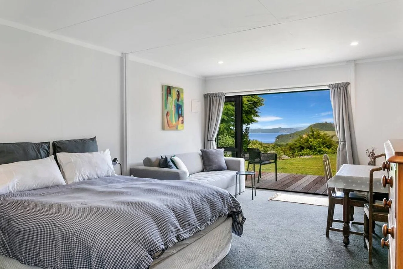 Room with a bed, gray checkered comforter, white pillows, a sofa with cushions, a painting on the wall, large window with a scenic view of greenery, water, and mountains, and a small balcony with outdoor chairs.