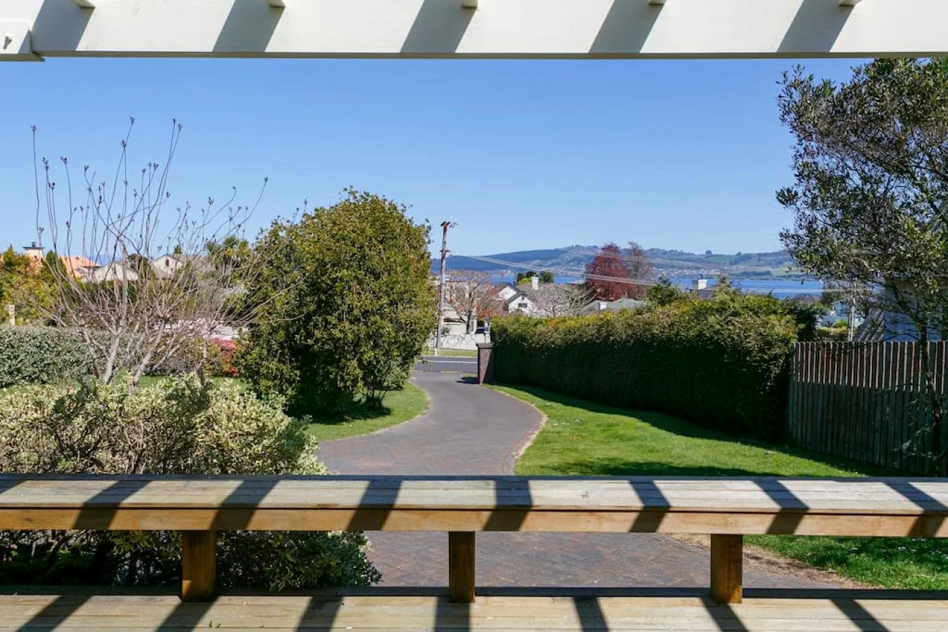 View from a porch overlooking a winding driveway, trees, shrubs, a fence, and a body of water in the distance.