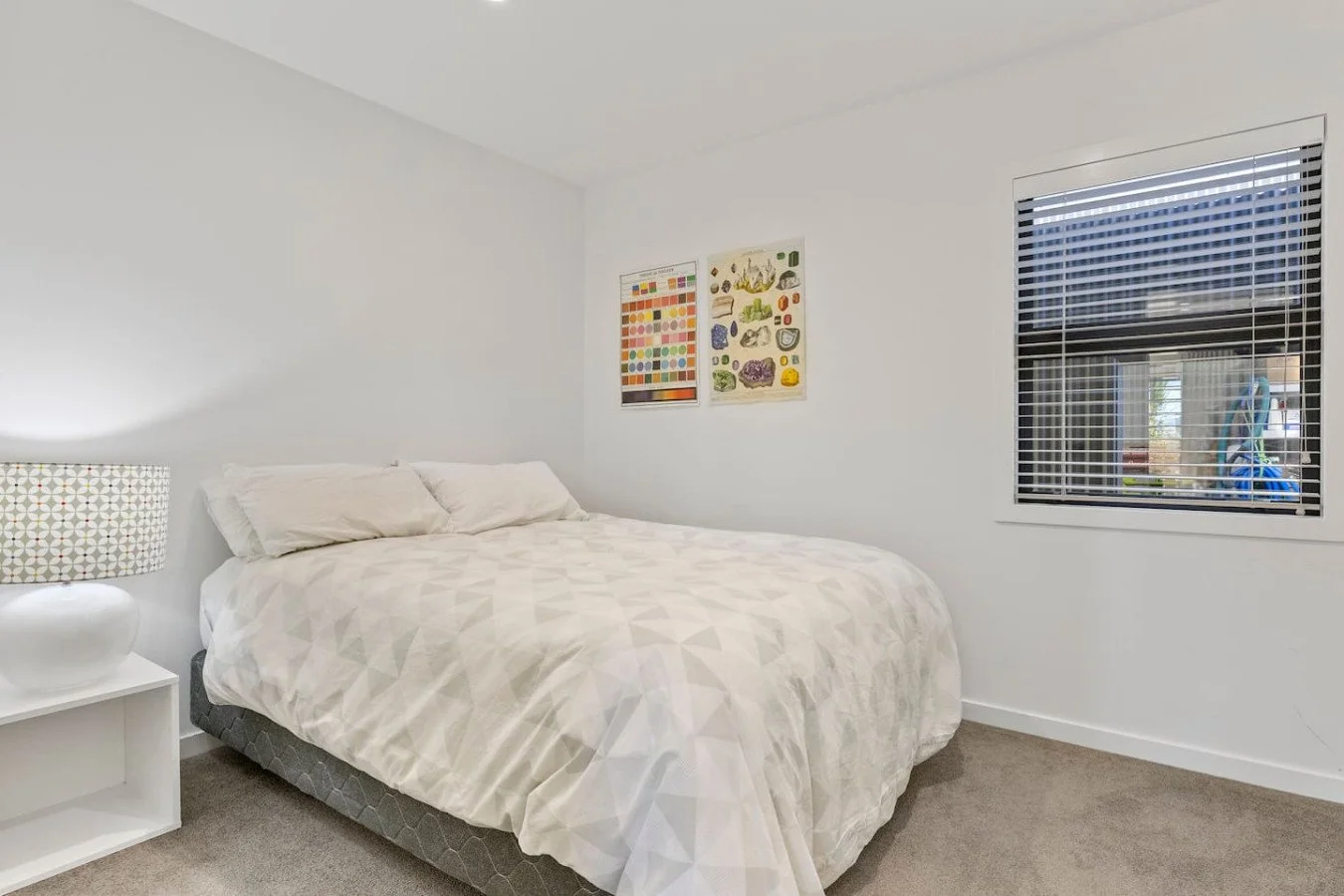 A bedroom with white walls, a bed with white bedding and pillows, a white nightstand with a patterned lamp, framed prints on the wall, and a window with blinds showing a view outside.